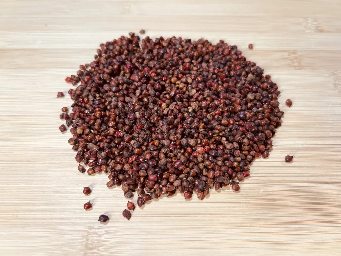 A small pile of cleaned winged sumac seeds, dark red-brown and round, on a light wooden cutting board